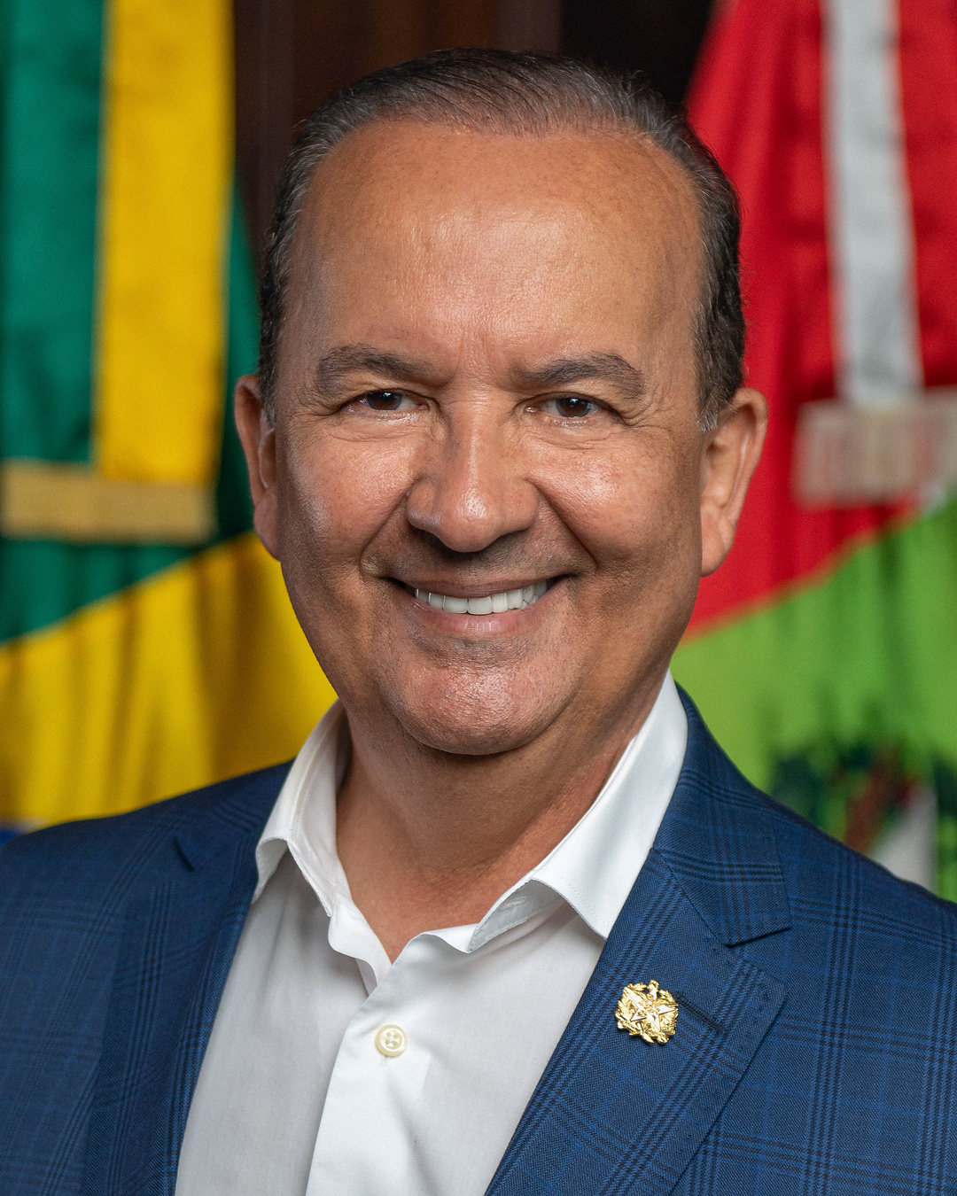 Retrato oficial de Jorginho Mello de paletó azul diante de bandeiras