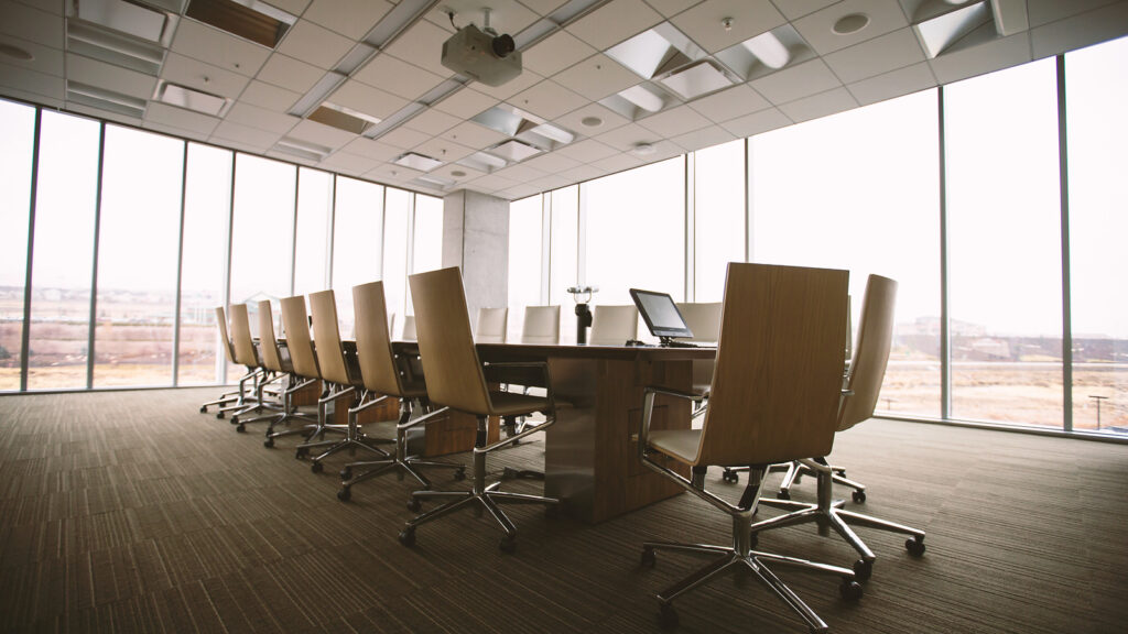 Sala de reunião vazia com mesa, cadeiras e janelas amplas.